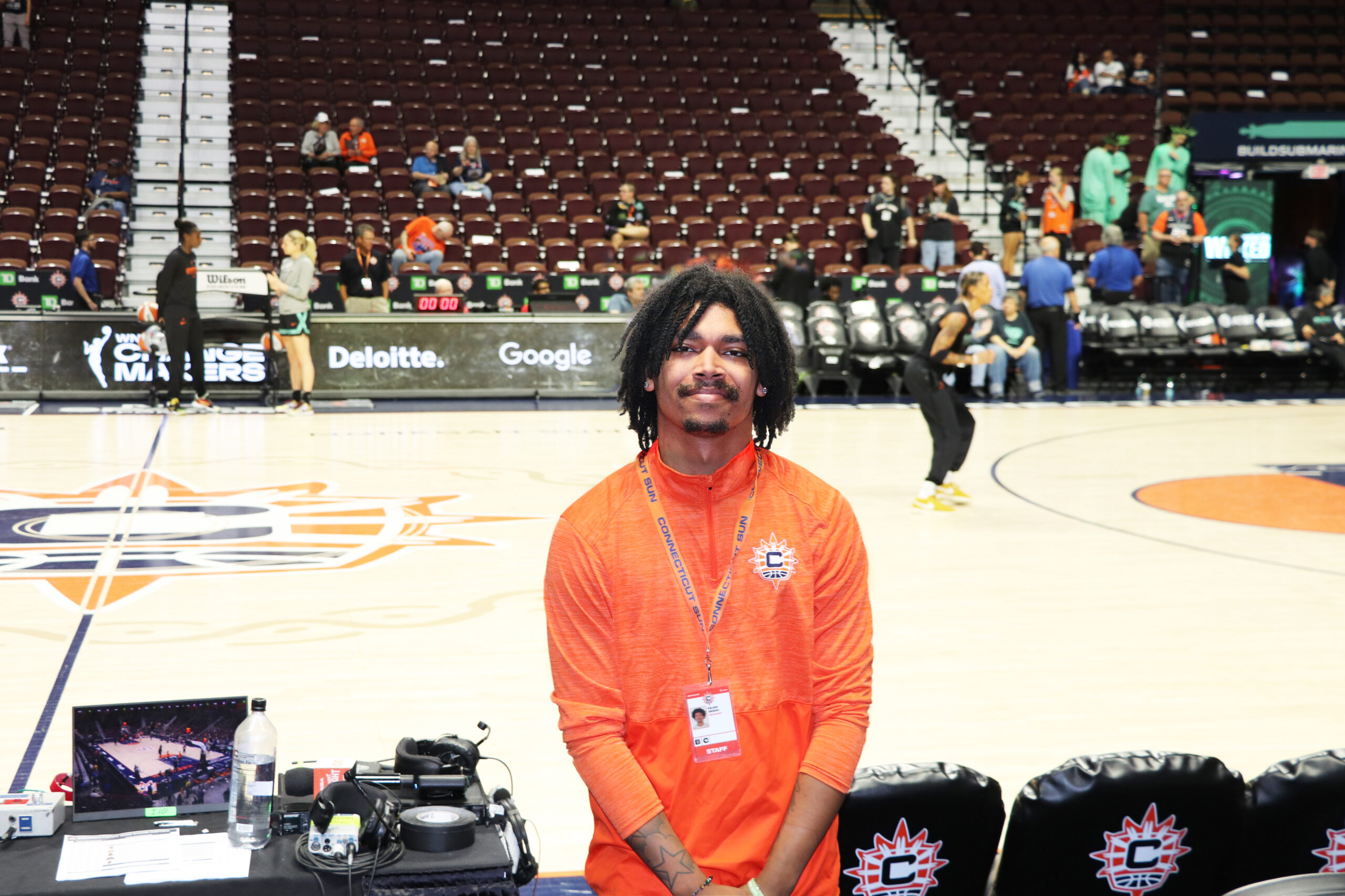 College student wearing orange Connecticut Sun shirt stands in front of basketball court as team warms up on gameday.